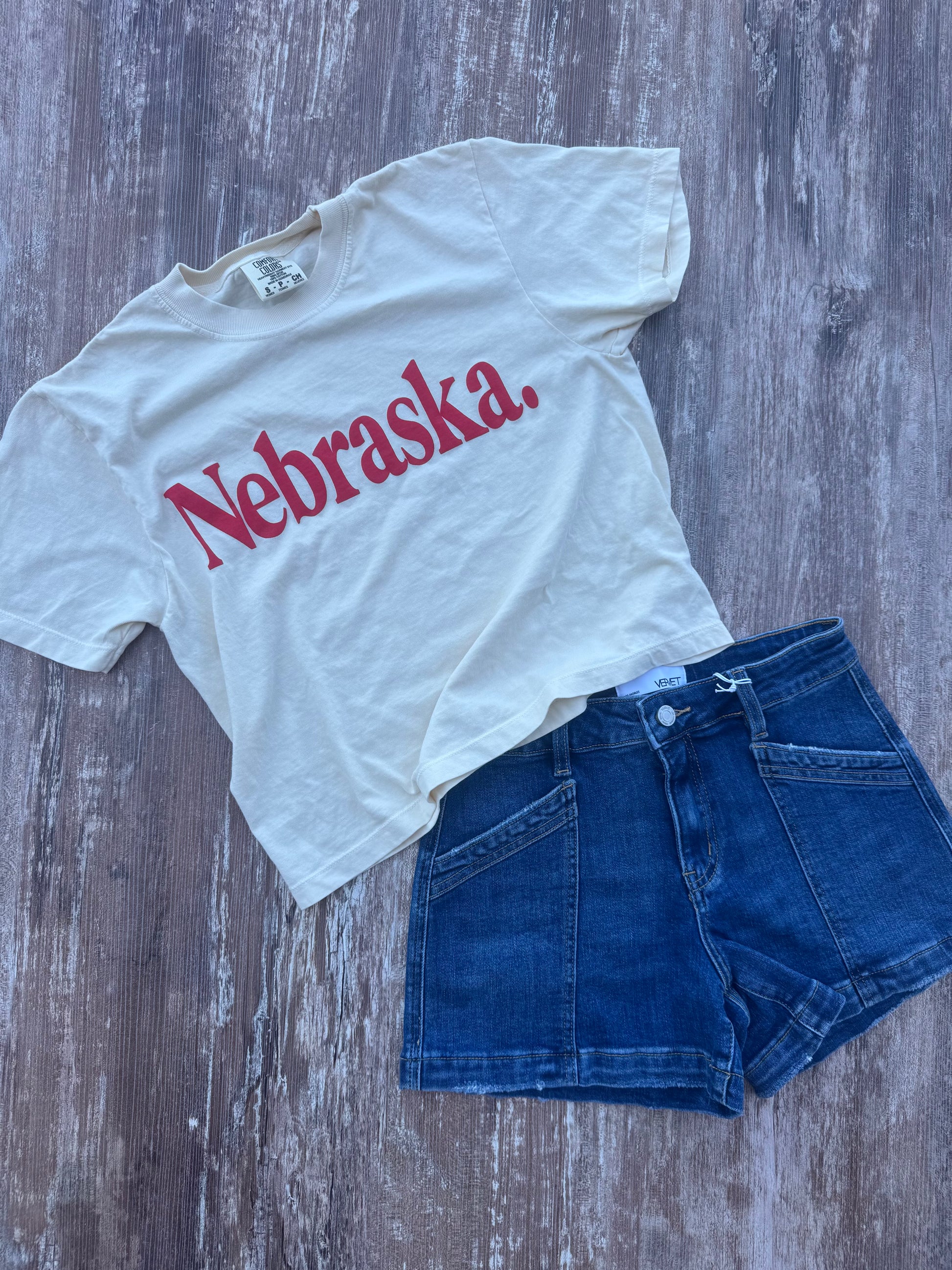 White t-shirt with 'Nebraska' printed in red and blue shorts on a wooden surface